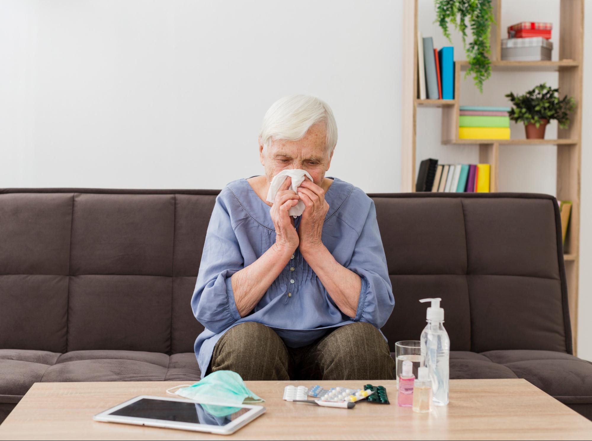 Elderly woman sneezing and appearing to have severe sinus pain while sitting at home, showing symptoms that may need care at Express Medical Clinic.

