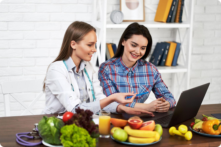 Doctor and patient at a medical weight loss clinic reviewing a diet chart on a laptop with fruits, salads, and weights on the table.