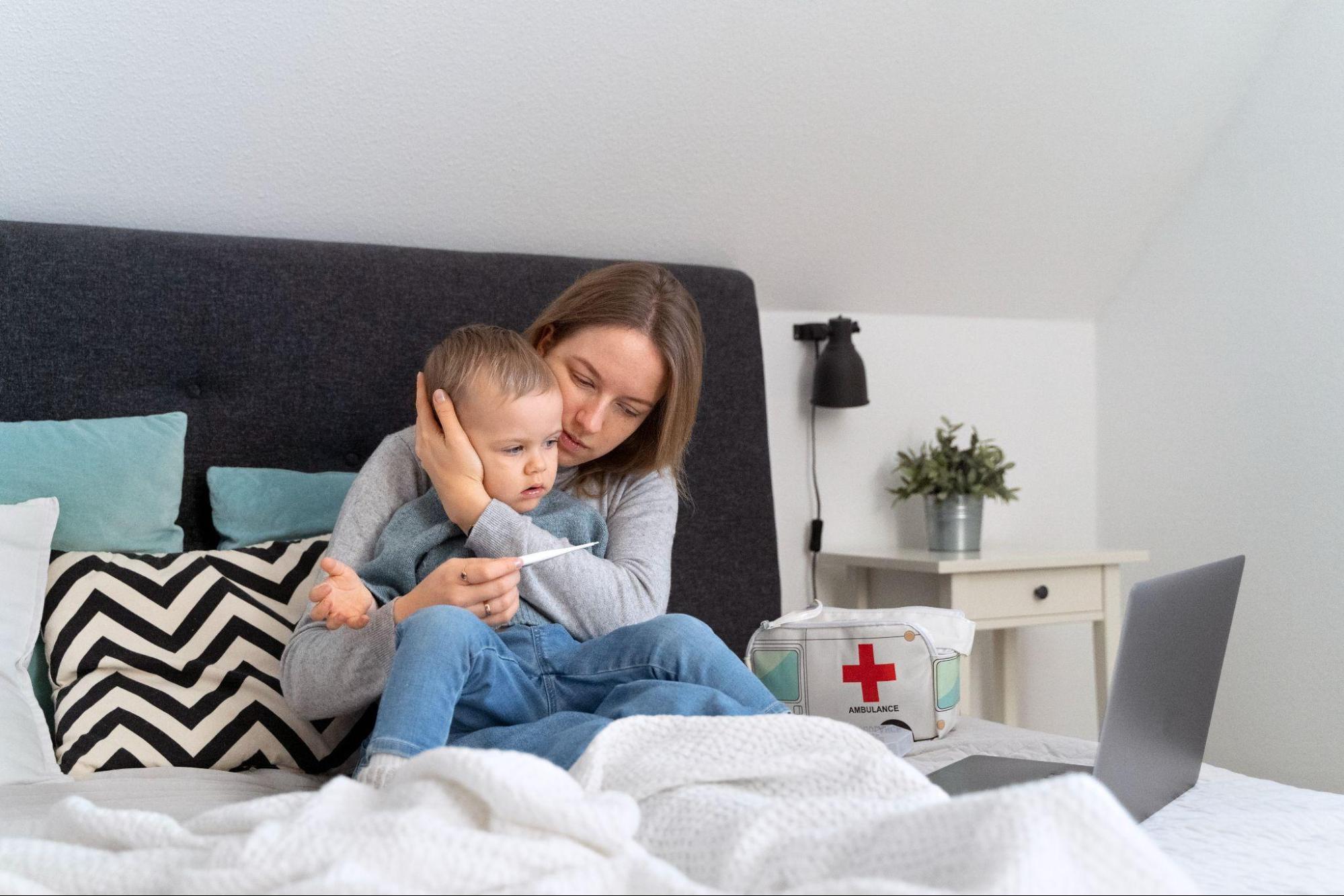 Mother hugs her baby boy while checking a thermometer, with a medicine box and laptop beside her — caring for her child with support from pediatric urgent care.